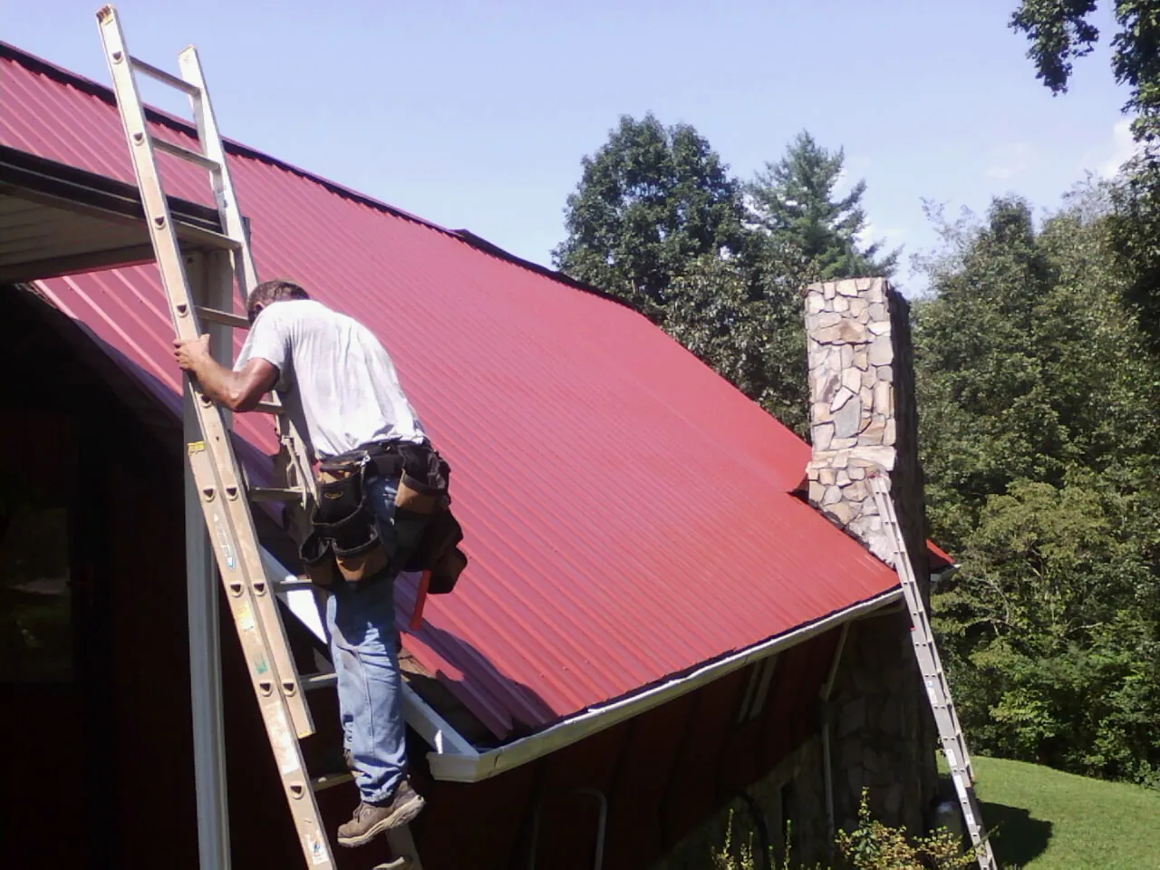 Roofer on a ladder beside a red metal roof and stone chimney — Soffit & Fascia Repair by Roofing Brothers in Massey